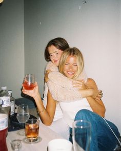 two women hugging each other while sitting at a table with drinks and glasses on it