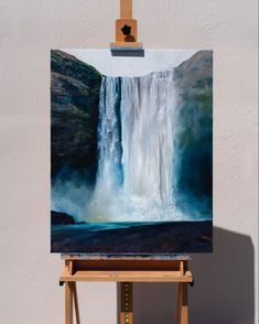 an easel with a painting on it in front of a white wall and a waterfall