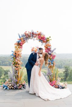 a bride and groom kissing in front of an arch with colorful flowers on it at their wedding