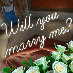 a bouquet of white roses sitting next to a neon sign that says will you marry me?