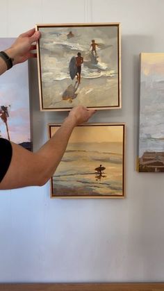 a man is holding up some paintings on the wall and pointing to them with his hand