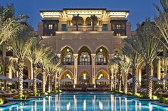 an outdoor swimming pool in front of a large building with palm trees and lights around it