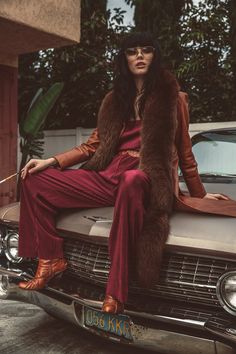 a woman sitting on top of a car wearing a fur coat and red pants with her legs crossed