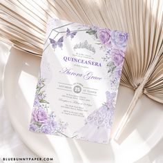 a wedding card with purple flowers on it next to an open fan and palm frond