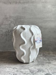 a white vase sitting on top of a cement floor next to a wall with a tag hanging from it's side