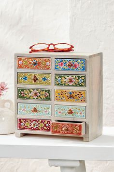 a multicolored wooden drawer with glasses on top and flowers in vase next to it