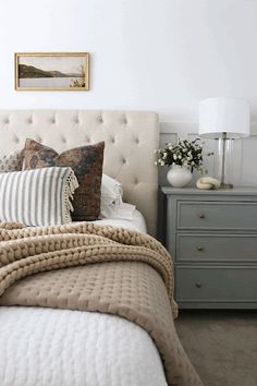 a bed with pillows and blankets on it in a bedroom next to a dresser, lamp and painting