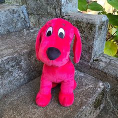 a pink dog stuffed animal sitting on top of a stone wall next to green leaves