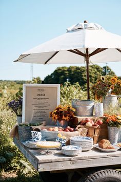 an outdoor table with flowers, fruit and plates on it in front of a white umbrella