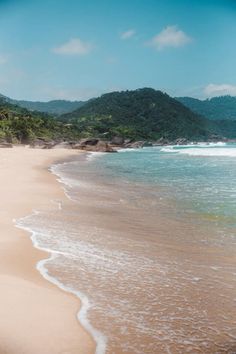 a sandy beach with waves coming in to the shore and green mountains in the background