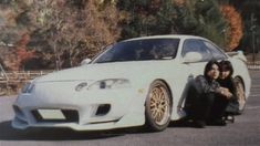 two people sitting next to a white car in a parking lot with trees behind them