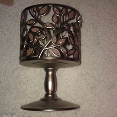 a silver candle holder with a tree design on the front and bottom, sitting on a white carpeted floor