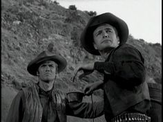 two men in cowboy hats are standing on the side of a hill with their arms stretched out