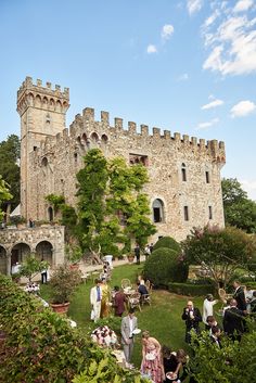 people are gathered in front of an old castle like building with trees and bushes surrounding it