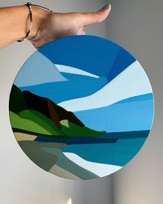 a hand is holding a circular painting in front of a white wall with blue sky and green hills