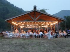 a group of people sitting at tables under a wooden structure with lights strung from it