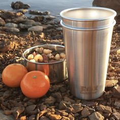 two oranges sitting next to a metal cup on the ground near rocks and water