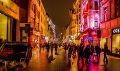 people walking down the street at night in an urban area with tall buildings and colorful lights