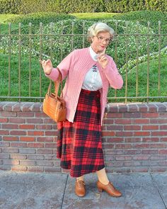 an older woman standing in front of a brick wall wearing a pink sweater and plaid skirt