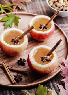 two apples with cinnamon sticks in them on a wooden plate next to nuts and leaves