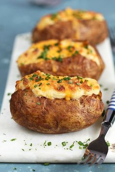 three baked potatoes with cheese and chives on a white plate next to a fork