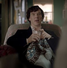 a young man sitting in a chair while holding scissors and knitting something to someone else