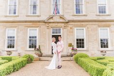 a bride and groom standing in front of a large building with hedges on both sides