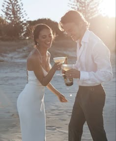 a man and woman standing next to each other on a beach holding wine glasses in their hands