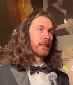 a man with long hair wearing a tuxedo and holding a microphone in front of him