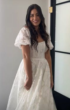 a woman in a white dress standing next to a door and smiling at the camera