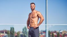 a shirtless man standing on the beach with his hands in his pockets and looking at the camera