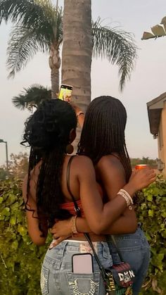 two women standing next to each other in front of a palm tree and looking at their cell phones