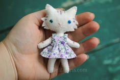 a small white cat doll in a dress on someone's hand with green background