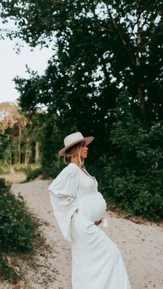 a pregnant woman in a white dress and hat is standing on a dirt road with trees behind her