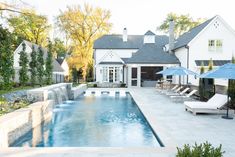 an outdoor pool with lounge chairs and umbrellas next to the swimming pool in front of a white house