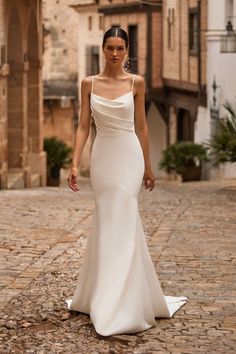 a woman in a white wedding dress walking down a cobblestone street with buildings behind her