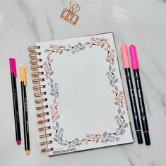 an open notebook with three pens next to it on a marble surface, surrounded by other stationery items