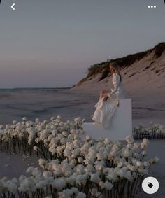 two women in white dresses are sitting on a small structure with flowers growing out of it