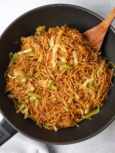 a wok filled with noodles and vegetables on top of a white table cloth next to a wooden spoon