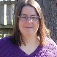 a woman wearing glasses standing in front of a tree with a fence behind her and looking at the camera