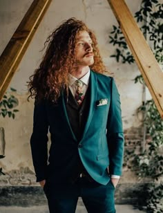 a man with long red hair wearing a blue suit and tie standing in front of a plant