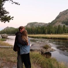 a man and woman standing next to each other in front of a river with mountains behind them