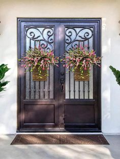 two flower baskets on the front door of a house with wrought iron doors and windows