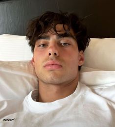 a man laying in bed with his eyes closed and looking at the camera while wearing a white t - shirt