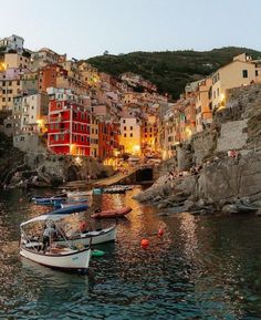 several small boats are docked in the water near some buildings and cliffs at night time