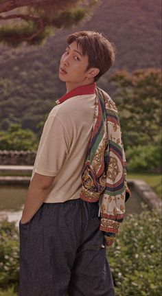 a young man with a backpack standing in front of some bushes and trees, looking off into the distance