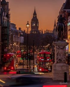 two red double decker buses driving down a street next to tall buildings and a clock tower