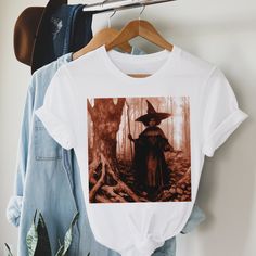 This adult unisex short sleeve tee features a dark and spooky tinted image of an afro witch in the woods. Crafted with soft cotton and a quality print, this tee will become your well-loved fall favorite. It's a great top to celebrate the spirit of Halloween, but the illustration works year round as well. Its ribbed knit collars, taping on the shoulders, and dual side seams ensure a better fit over time and maintain the garment's shape. Made with 100% cotton, this tee offers a comfortable retail Grunge T-shirt For Fall Fan Merchandise, Unisex Fall Short Sleeve T-shirt, Halloween Cotton Band Merch Tops, Cotton Halloween Band Merch Tops, Unisex Graphic Print T-shirt For Fall, Cotton Band Merch Tops For Halloween, Unisex Short Sleeve T-shirt For Fall, Fitted Band Merch T-shirt For Fall, Fall Short Sleeve T-shirt