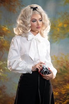 A blond pinup model posing against a yellow and blue painted background while wearing the Angela Blouse in white and black trousers with an old camera on her hand. Fitted White Blouse With Bow, Fall Puff Sleeve Blouse With Bow, Fall Puff Sleeve Blouse With Bow Detail, Classic Formal Blouse With Balloon Sleeves, Fall White Blouse With Tie Sleeves, Classic White Satin Blouse, White Tie Sleeve Blouse For Fall, White Tie Sleeves Blouse For Fall, White Fitted Tie Neck Blouse