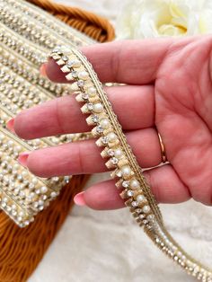 a woman's hand holding a gold and white beaded bracelet with flowers in the background